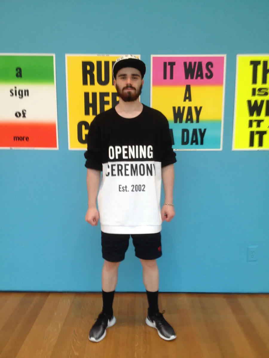 A young man in a cap and Opening Ceremony sweatshirt stands in front of bright text posters on a blue wall, photograph by Esther Piaskowski