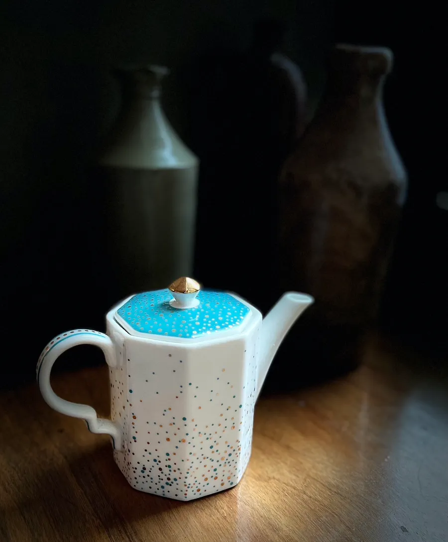 A white porcelain teapot with a turquoise lid on a dark wooden table, old bottles behind it in shadow, photograph by Esther Piaskowski